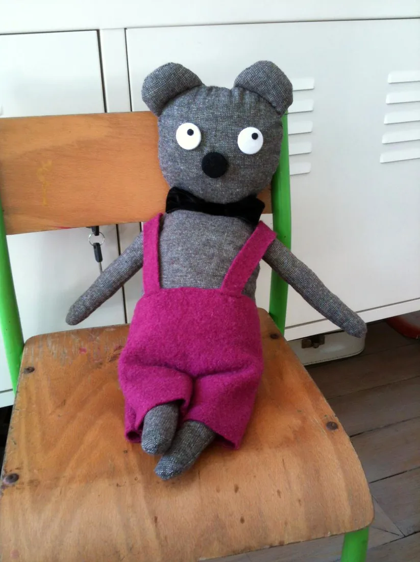 Un ours gris en salopette rose, assis sur une chaise d'écolier.